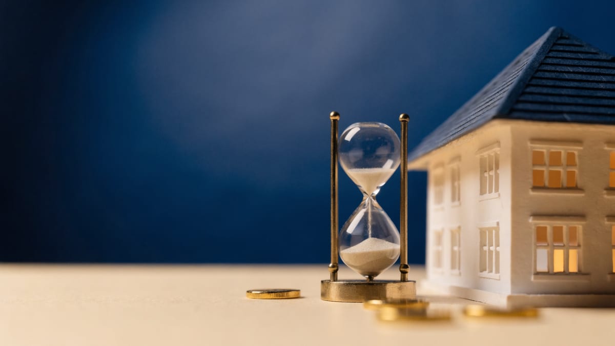 Scale model house beside a small brass hourglass on a navy backdrop, representing a fixed rate term ending